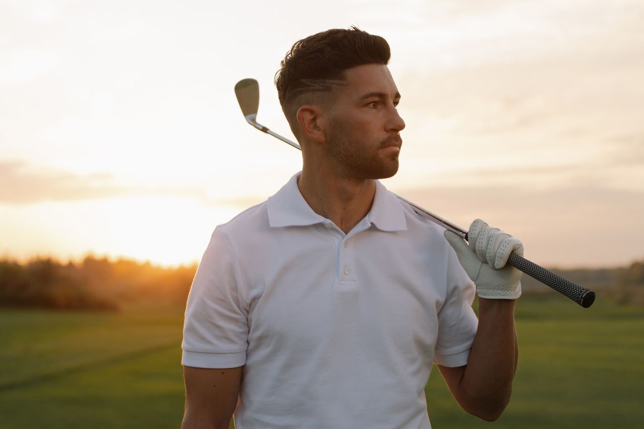 services-03 Portrait of a stylish male golfer posing with a club at sunset on a golf course, wearing a white polo shirt.