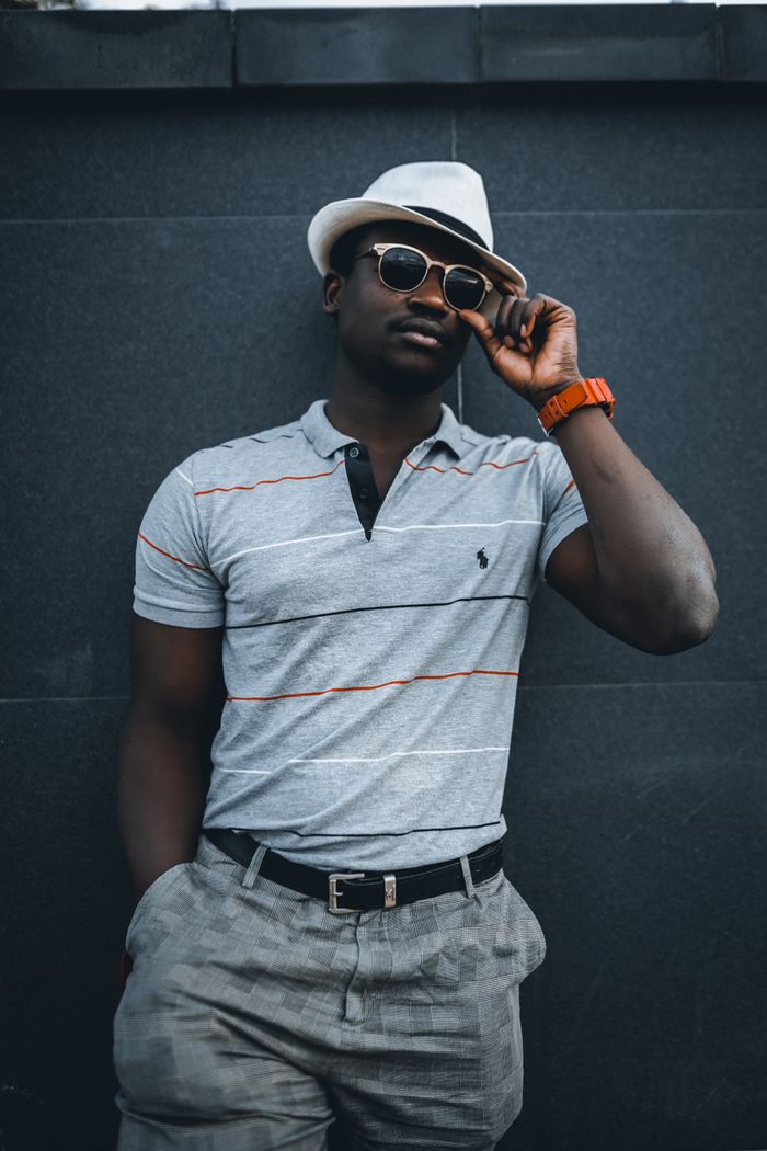 who-we-are Fashionable African American man wearing a hat and sunglasses, leaning casually for a stylish portrait.